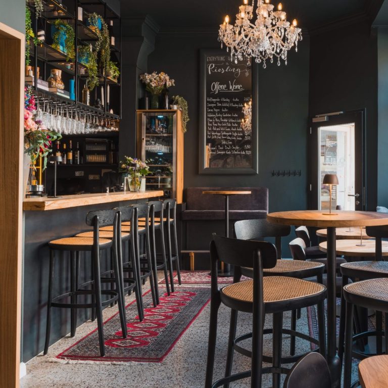 Moderne Bar mit Holzregal, Pflanzen, Kronleuchter und eleganten Sitzplätzen.